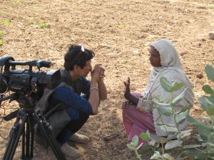 Reena crouches beside her camera to speak to a woman in a headscarf on dry, sunny ground.