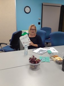 Brenda is shown sitting at the meeting table holding the book, with papers and meeting snacks in front of her.